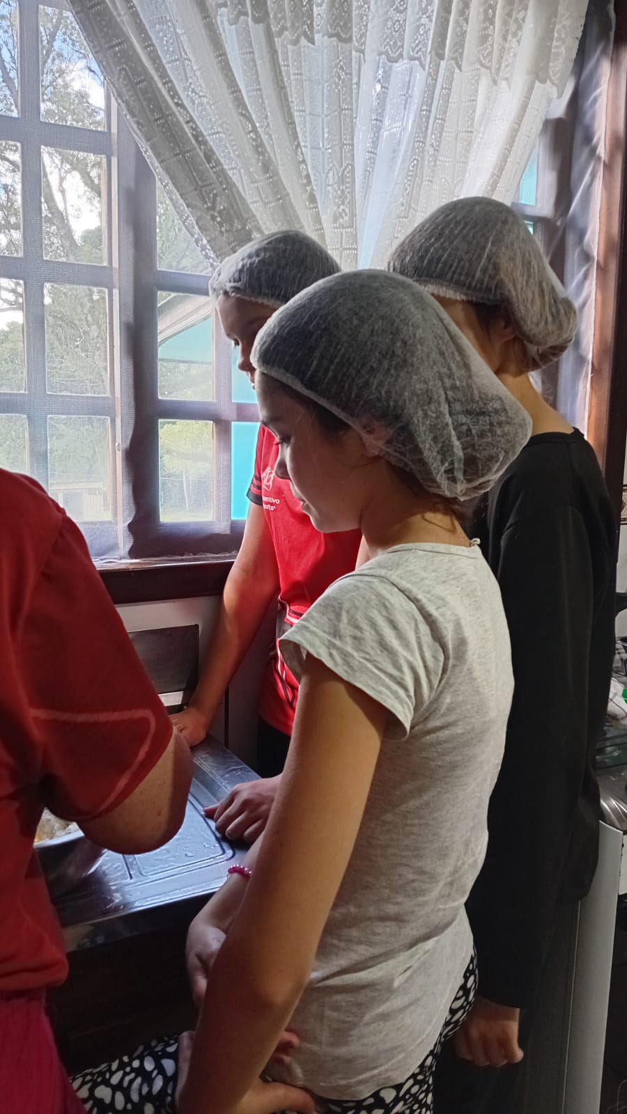 Três adolescentes de touca acompanham a voluntária preparando a massa na bancada da cozinha